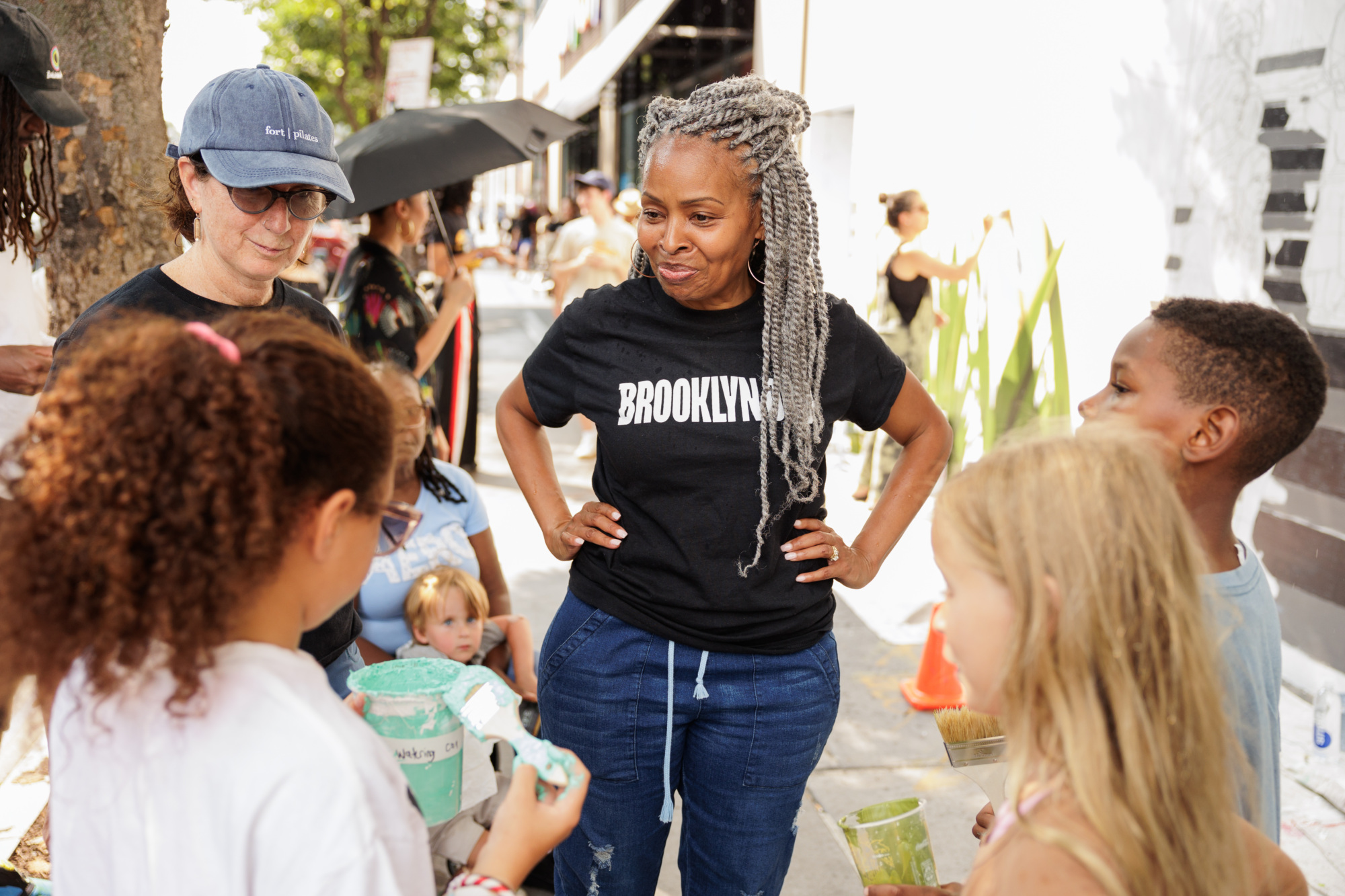 Woman with gray braids and "Brooklyn" t-shirt stands with hands on hips, surrounded by children holding paint. Outdoor setting with people in background.