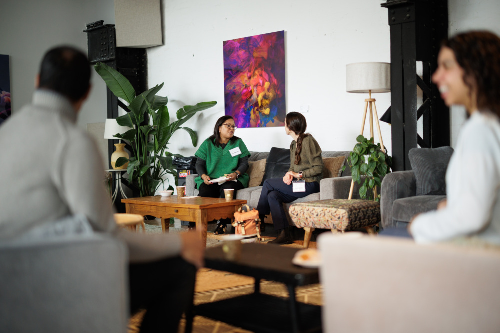Four people sit in a modern living room; two in conversation at the center, others are blurred in the foreground on either side. A colorful painting and plants decorate the space.