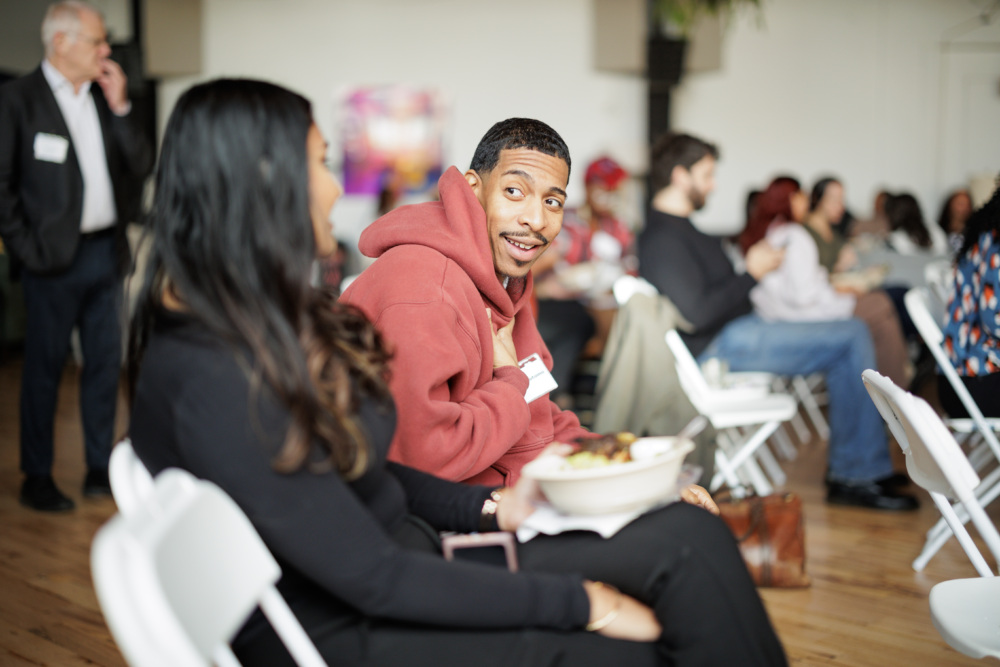 A man in a red hoodie gestures while talking to a woman with long hair, seated beside him in a room with others on white chairs.