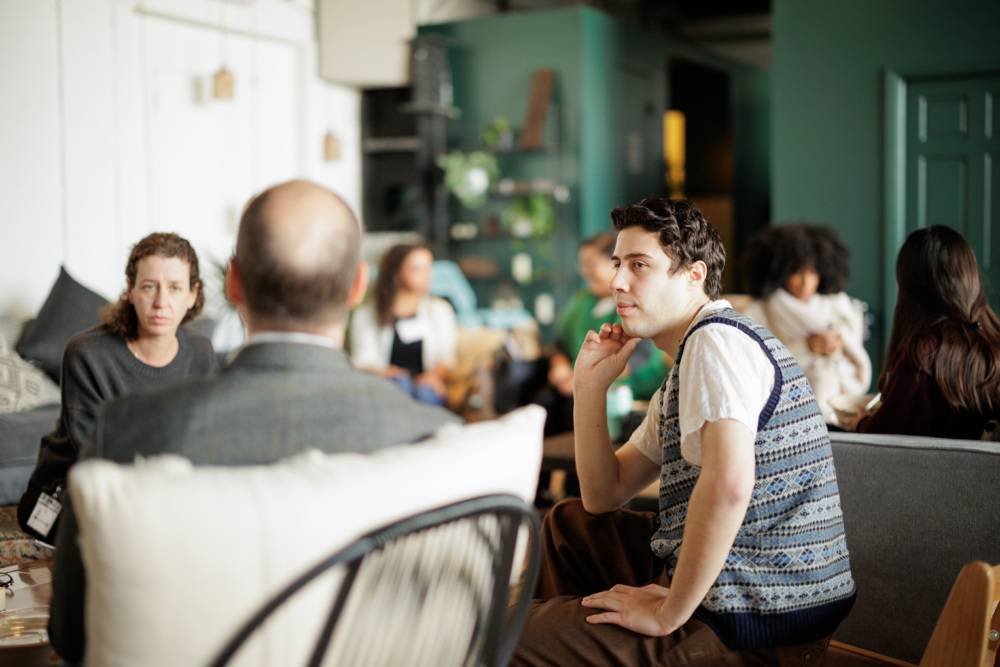 A group of people seated in a casual indoor space, engaged in conversation. A man in a patterned vest sits in the foreground.