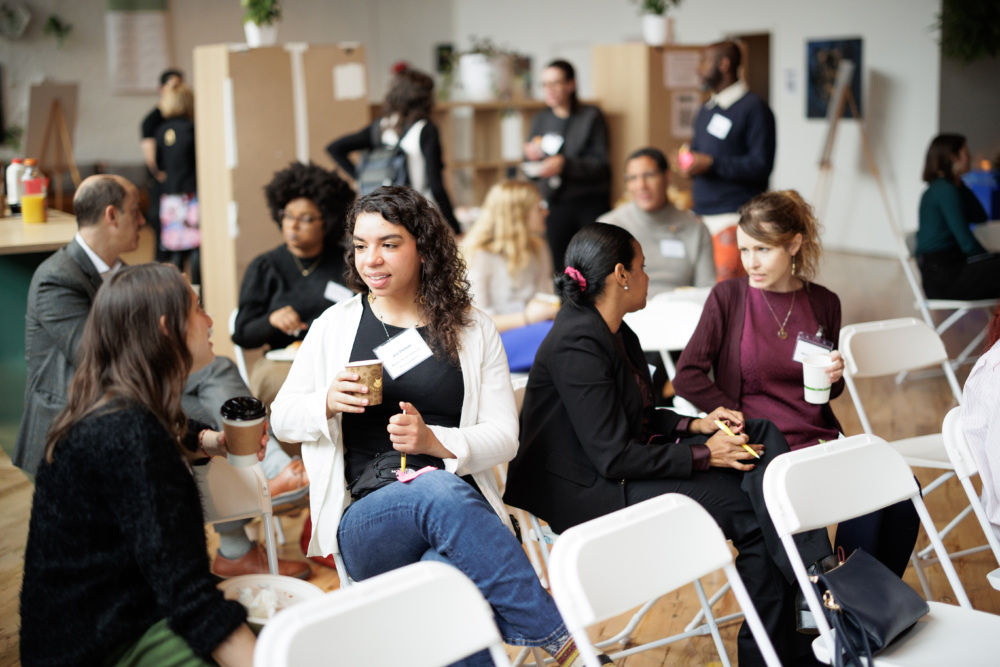 People sitting and talking in a casual indoor gathering, holding drinks and snacks. White folding chairs and tables with food are visible in the background.