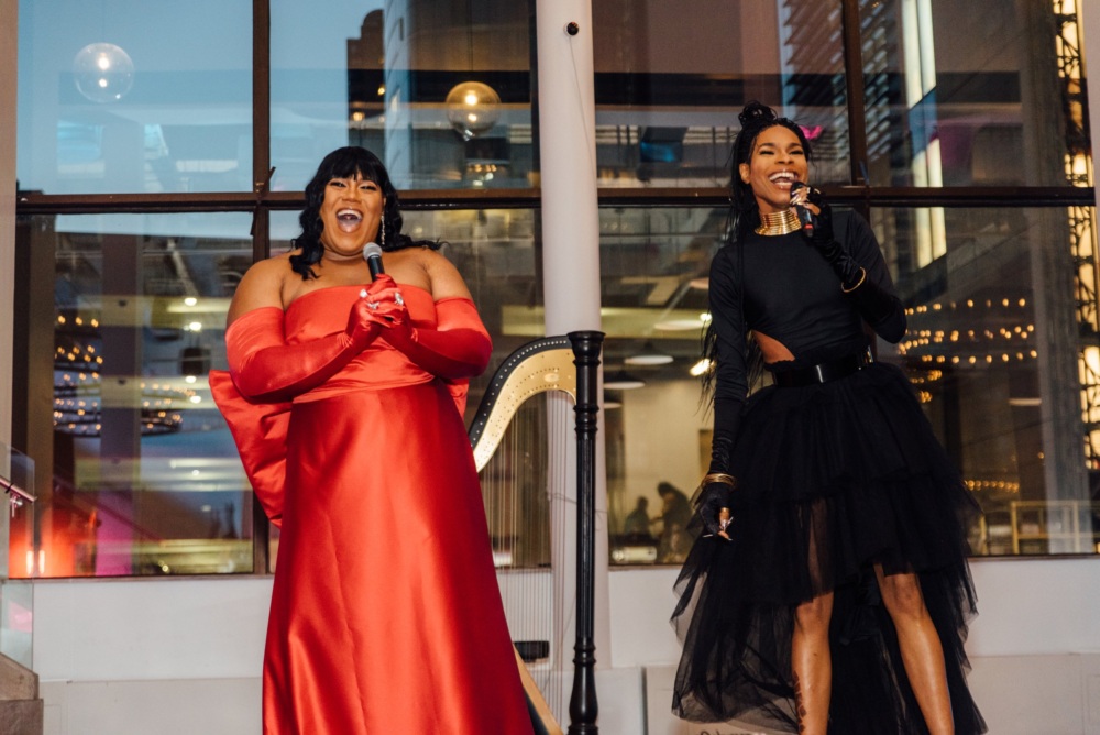 Two people holding microphones, smiling and singing. One wears a red gown, the other a black dress. They're indoors with a large window and city lights in the background.