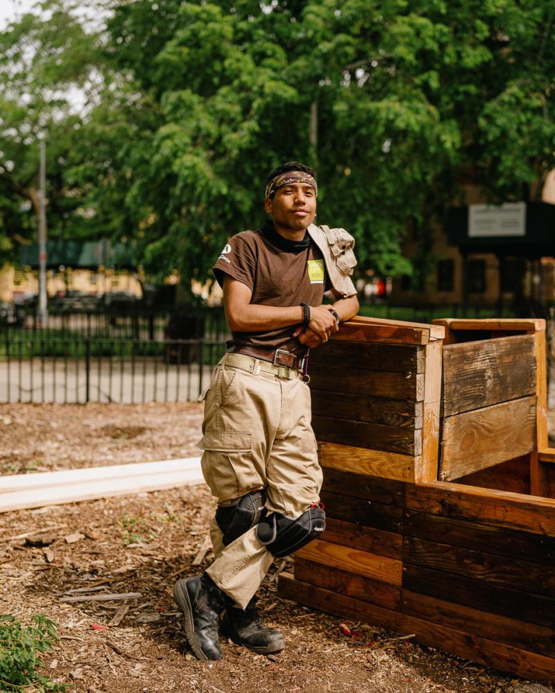 Person in work attire and knee pads leans against a wooden structure outdoors, with trees and a park in the background.