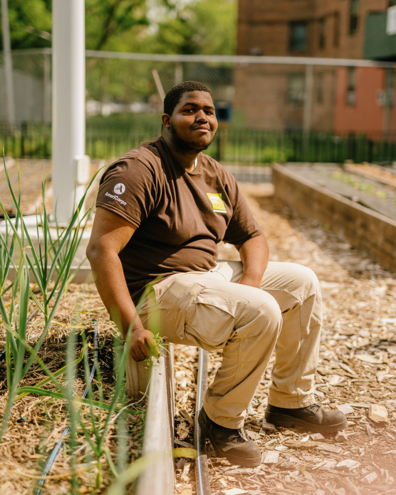 A person in a brown shirt and beige pants is sitting on a garden bed, surrounded by plants, with a building and fence in the background.