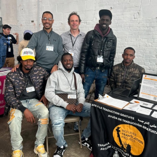 Six men pose together at a booth for the Refugee Translation Project, featuring information and promotional materials.