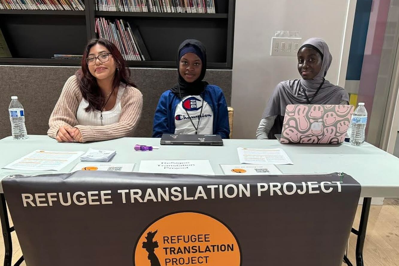 Three women sit at a table with a sign reading "Refugee Translation Project," offering translation support for asylum seekers and immigrants. The table has documents and water bottles.