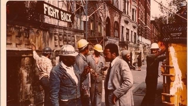 Workers in hard hats and casual clothing stand talking on a city street beside a "Fight Back" sign, with construction equipment in use nearby.