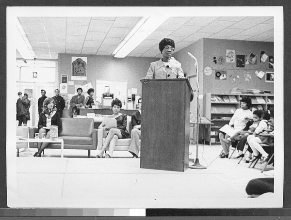 A woman speaks at a podium in a room with seated and standing audience members. Bookshelves and posters are visible in the background.