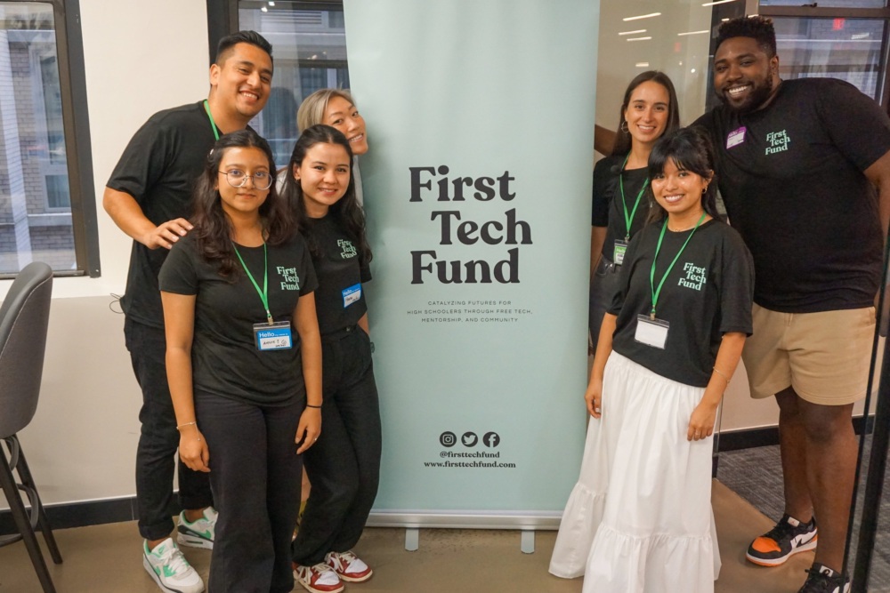 A group of seven people stands smiling in front of a "First Tech Fund" banner. They are wearing black shirts and have name tags.