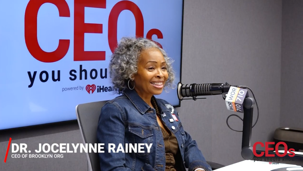 Woman with gray curly hair sits in a radio studio, smiling, wearing a denim jacket. A microphone is in front of her and a screen behind displays logo text with "CEOs You Should Know.