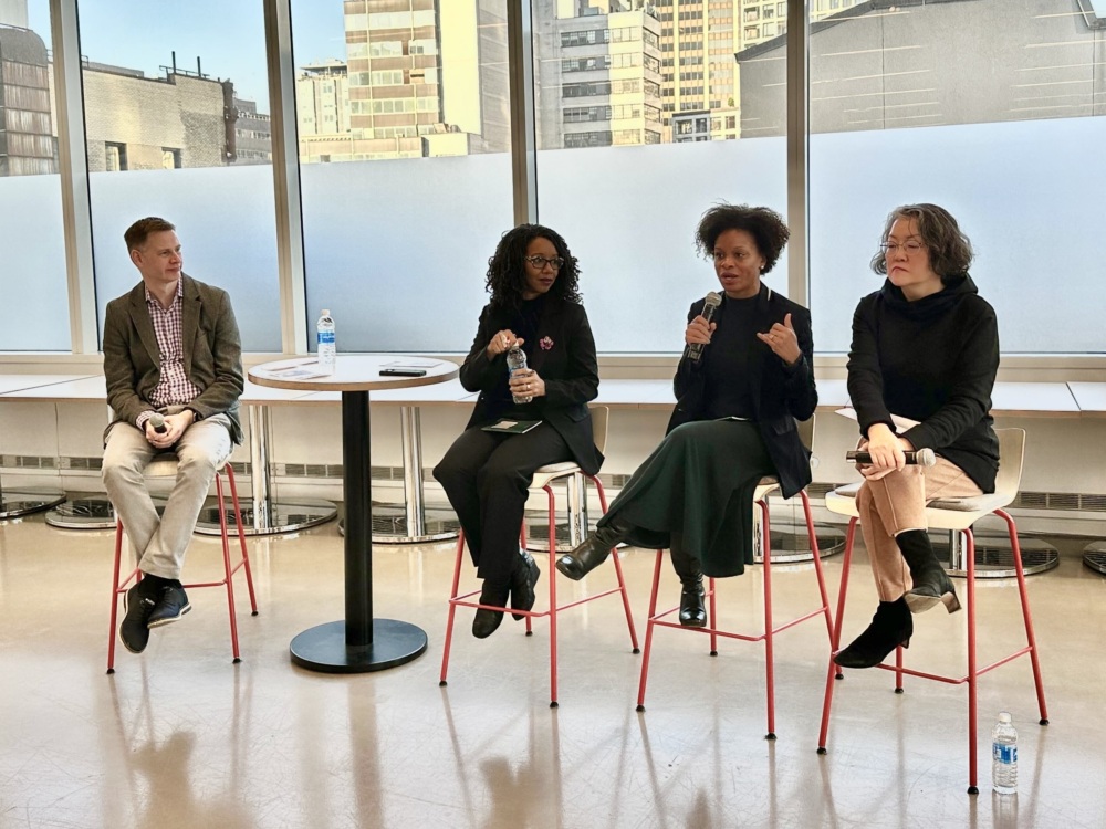 Four people sit on stools in a panel discussion setting with city buildings visible outside the windows.