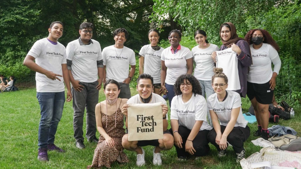 A group of thirteen people posing outdoors, all wearing "First Tech Fund" t-shirts. One person holds a sign with the same text. Trees and grass are in the background.