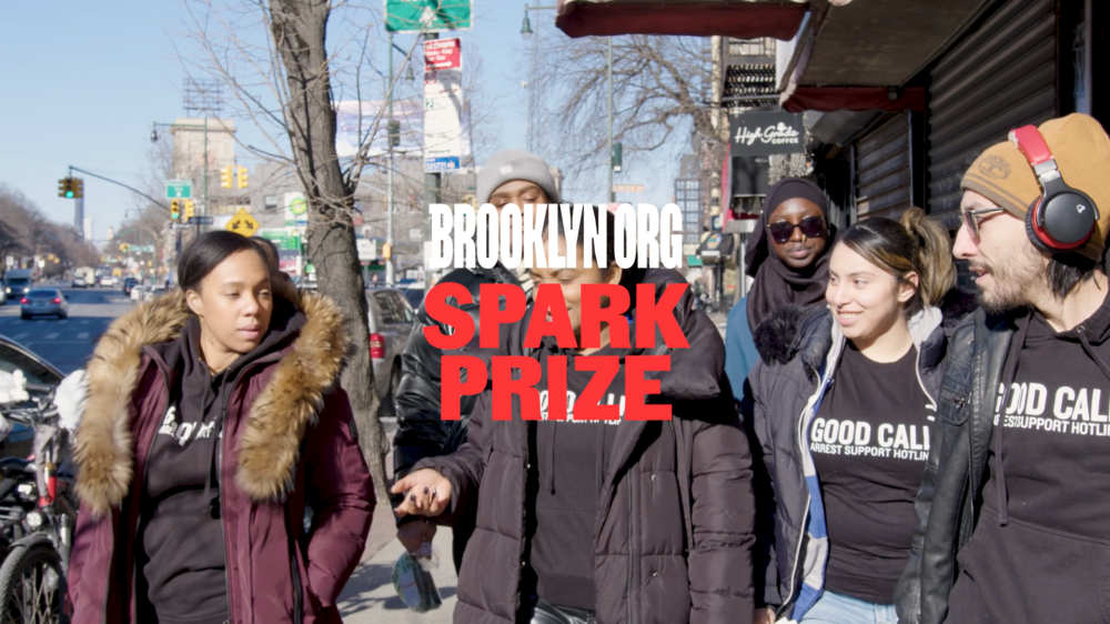 A group of people in winter clothing walk along a city street, with "Brooklyn.org Spark Prize" text overlayed at the center of the image.