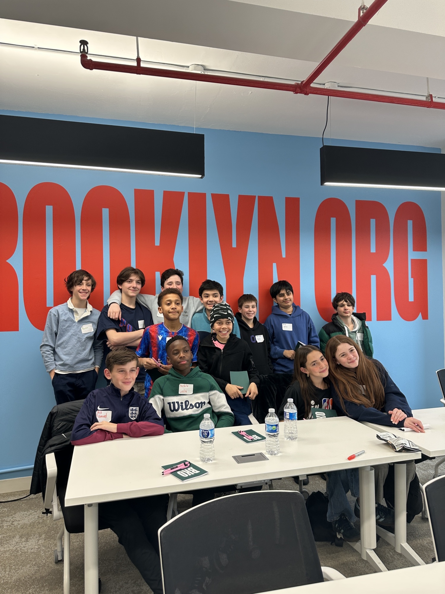 Group of 13 young people posing together in a room with a large sign reading "BROOKLYN.ORG" on the blue wall behind them.