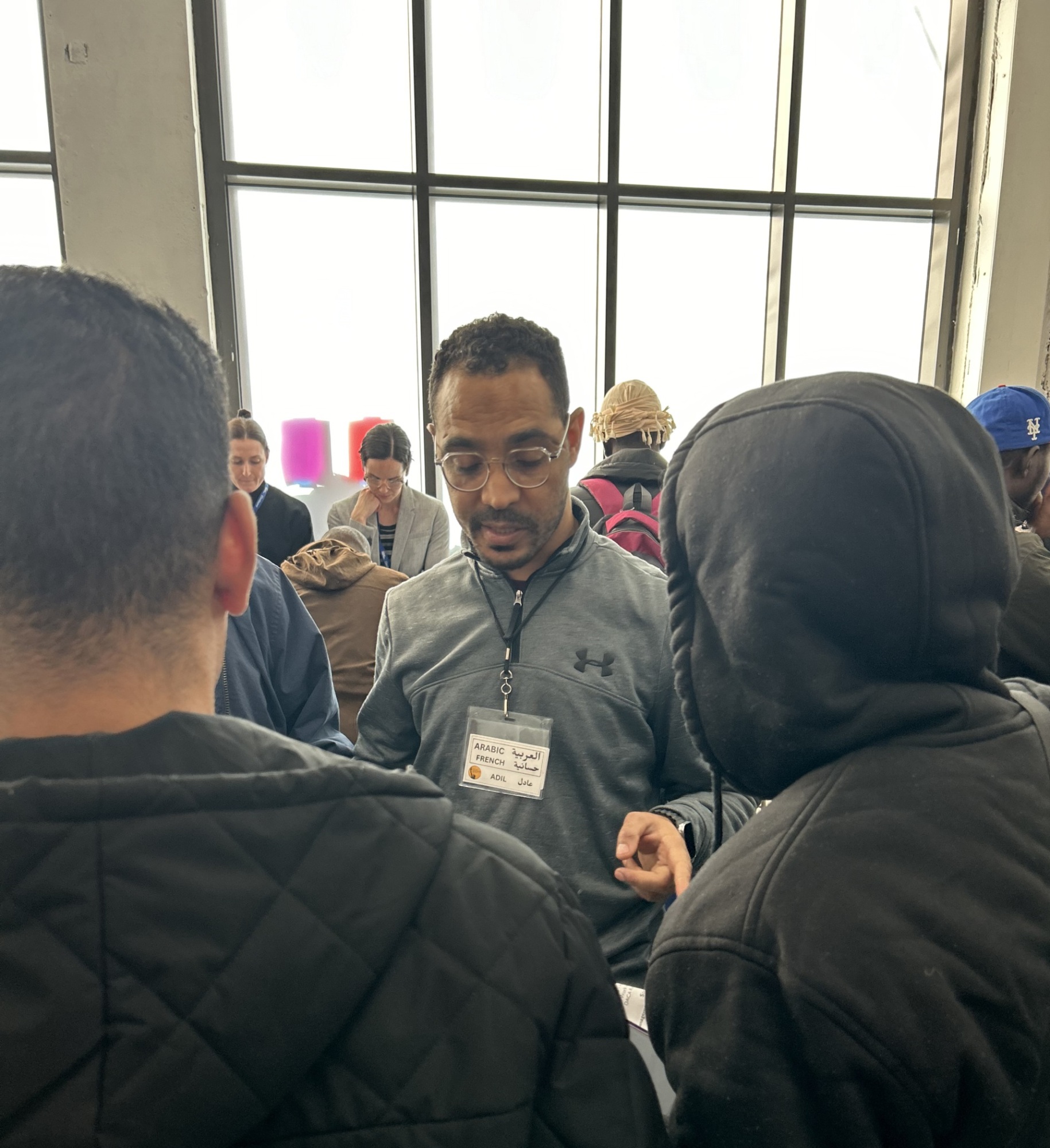 A man in glasses with a name tag talks to two people wearing hooded jackets in a busy indoor setting.