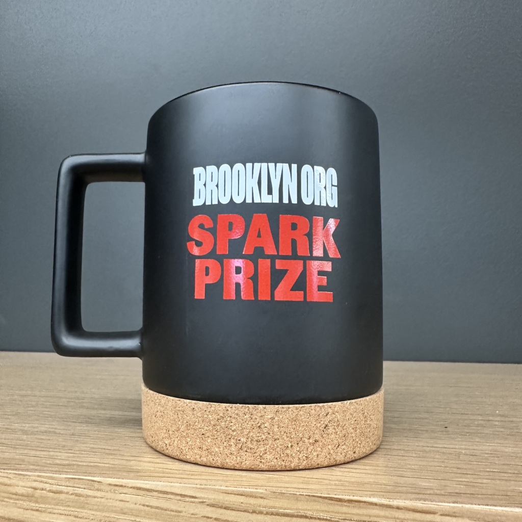 A black mug with "Brooklyn Org Spark Prize" printed in white and red letters. The bottom has a cork base.