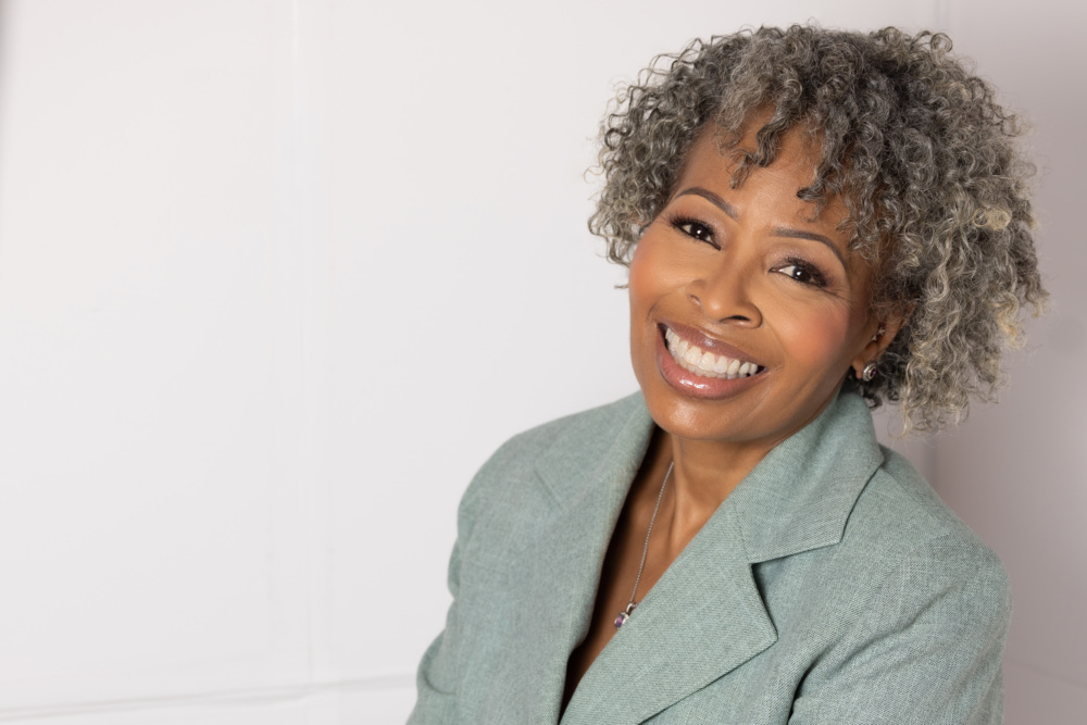 A person with curly gray hair smiling, wearing a light green blazer, and standing against a white background.