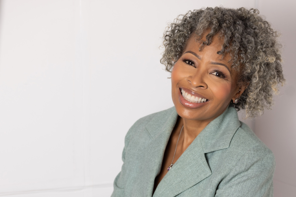 A person with curly gray hair smiling, wearing a light green blazer, and standing against a white background.