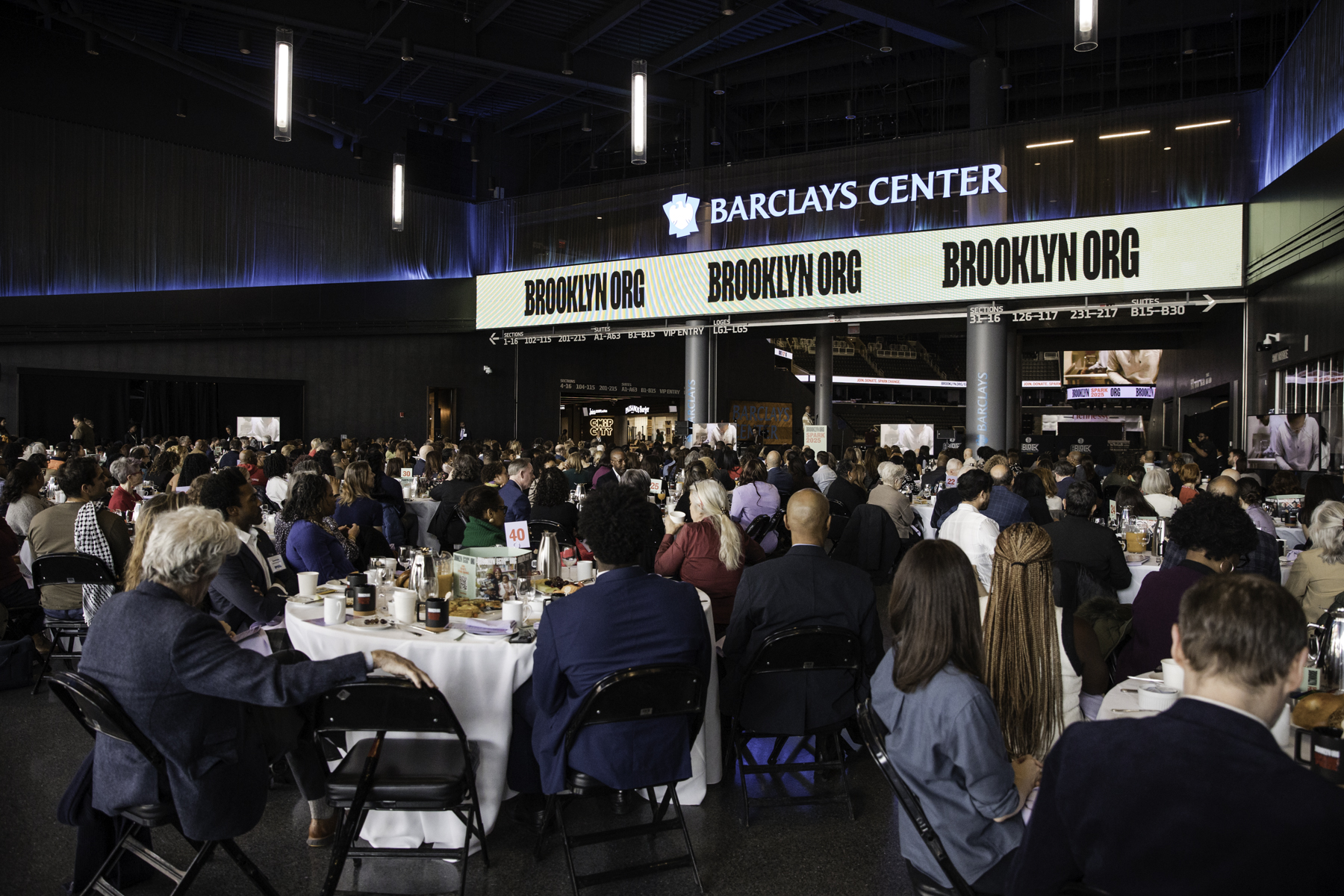 A large crowd attends an event at Barclays Center, sitting at round tables with food and drinks, under screens displaying "BROOKLYN" and Barclays Center logos.