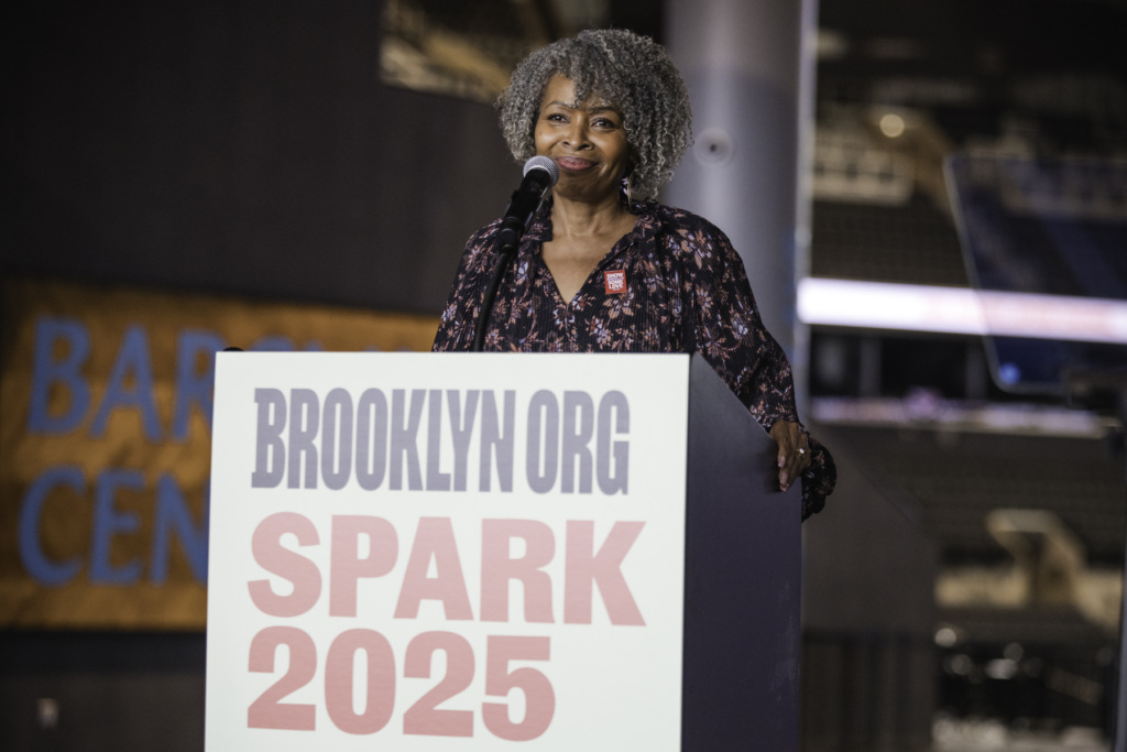 A person with gray curly hair stands at a podium with a microphone, speaking at an event labeled "BROOKLYN ORG SPARK 2025.