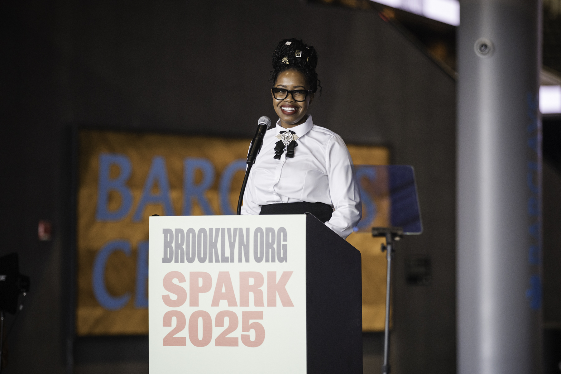 A person stands at a podium with a microphone, smiling, in front of a sign that reads "Brooklyn.org Spark 2025.