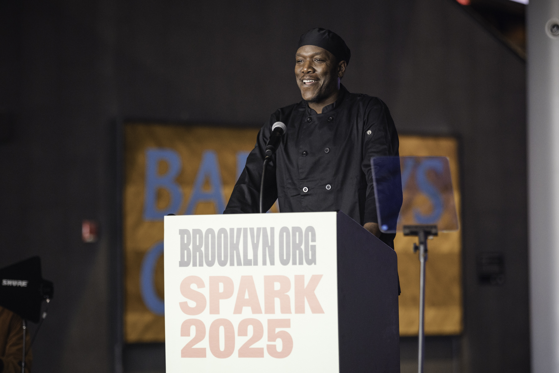 Person in a black chef uniform speaking at a podium labeled "Brooklyn.org Spark 2025," with a microphone and a background banner partially visible.