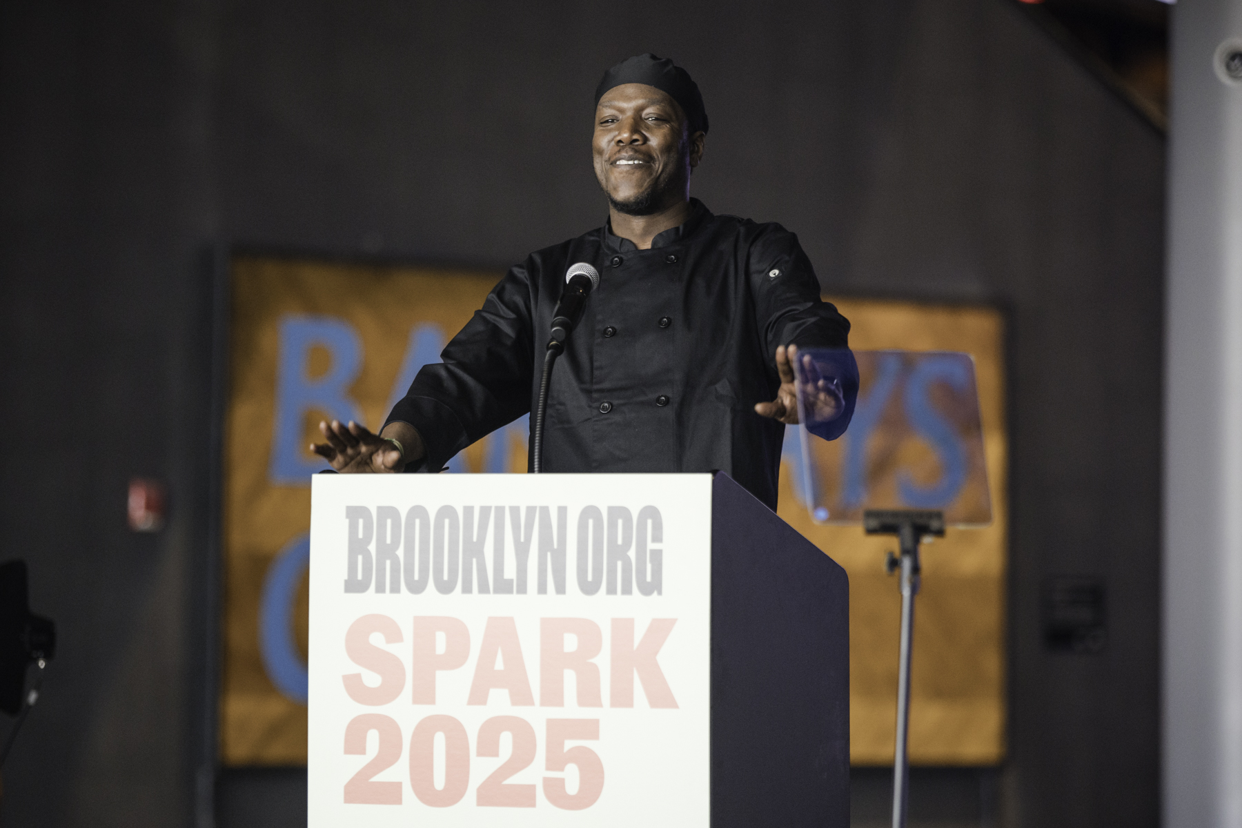 A person in a black chef's outfit stands at a podium labeled "BROOKLYN.ORG SPARK 2025," speaking into a microphone.