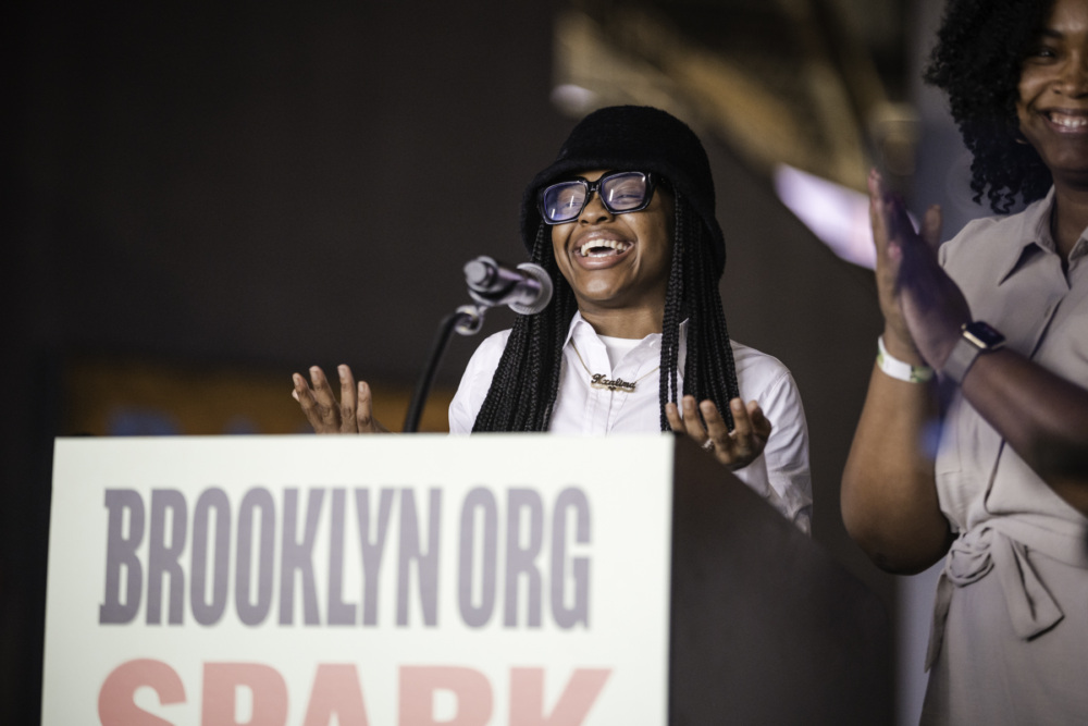 A person in glasses and a hat speaks and gestures at a podium with "BROOKLYN.ORG SPARK" on it, while another person claps nearby.