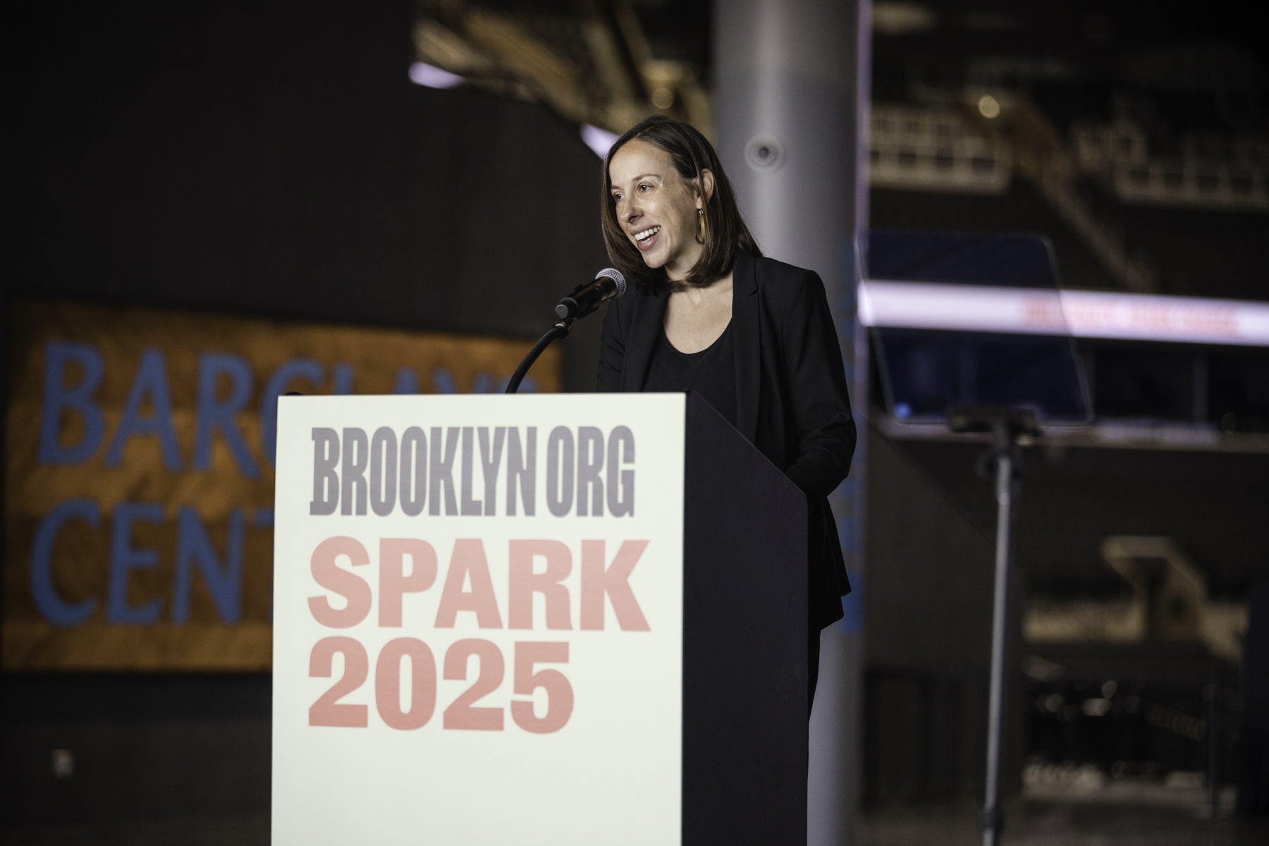 A person speaks at a podium with a "BROOKLYN.ORG SPARK 2025" sign. The venue is Barclay Center.