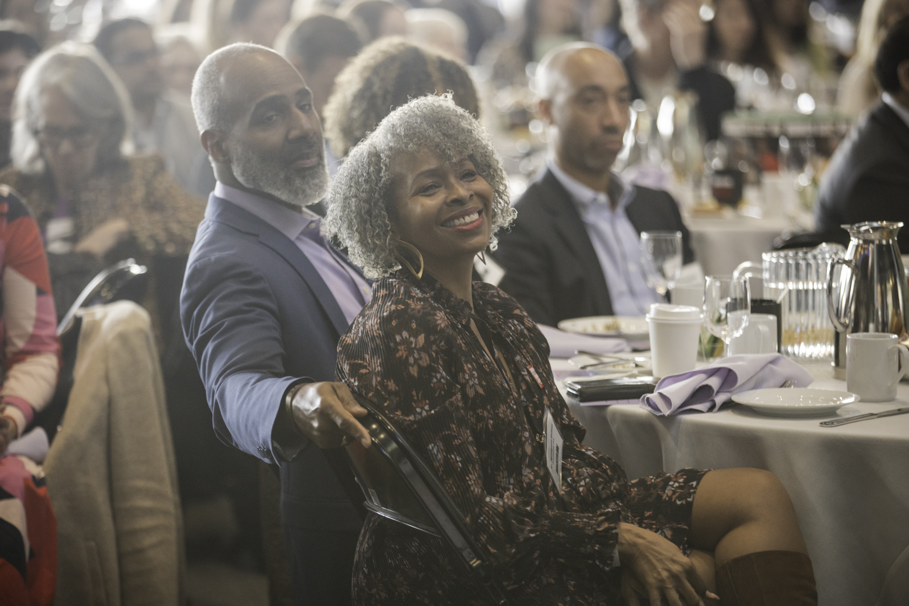 People seated at tables during an indoor event, with a focus on a smiling woman in a floral dress and a man in a suit jacket.