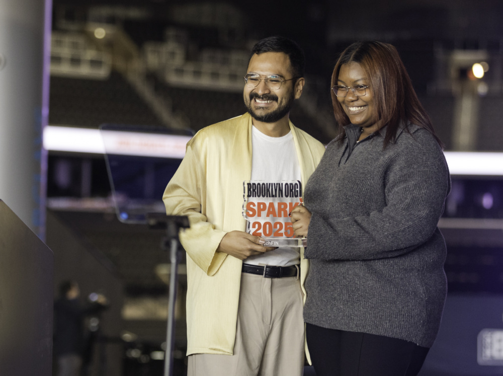 Two people smiling and posing for a photo on stage at a "Brooklyn Org Spark 2023" event, with a podium and microphone nearby.
