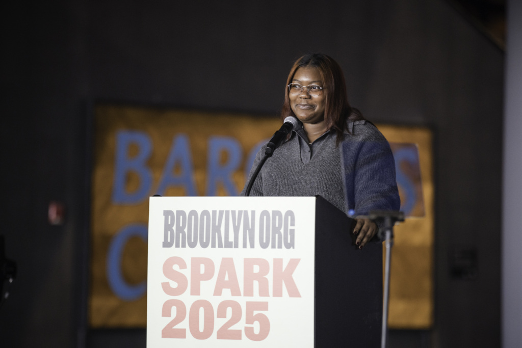 A woman speaks at a podium labeled "Brooklyn.org Spark 2025," with a blurred banner in the background.