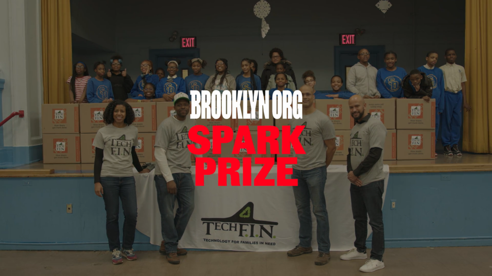 Group of people standing in front of a table with Tech FIN logo, boxes, and children on stage. Text reads "Brooklyn.org Spark Prize.