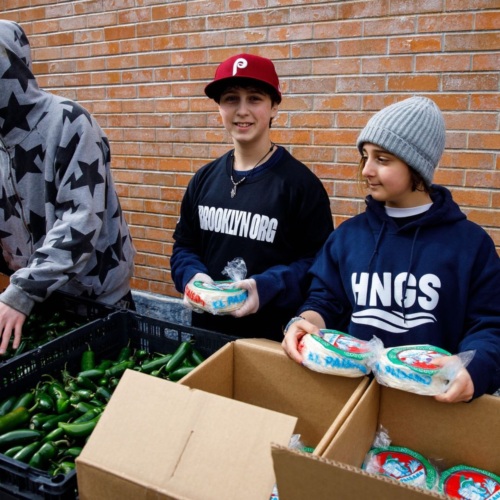 Three individuals organizing produce and packaged goods outdoors by a brick wall. Two are wearing hoodies and one a burgundy baseball cap.