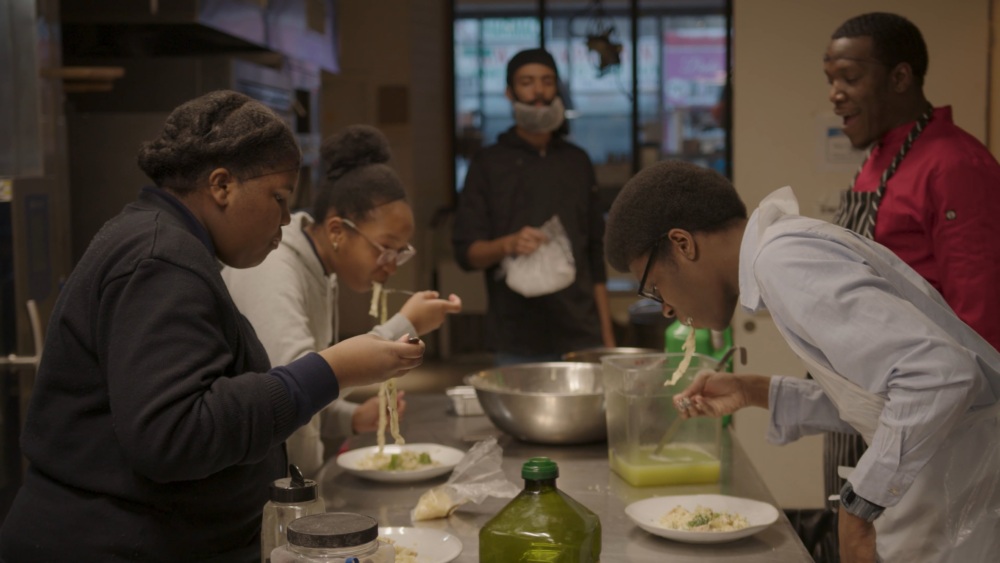 People gathered around a kitchen counter tasting food, with bowls, plates, and bottles visible. A person in the background wears a mask.