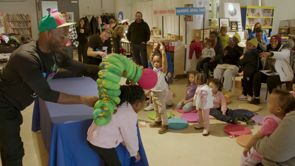 Performer with green caterpillar puppet entertains a group of young children sitting on colorful cushions in a community center, as adults watch in the background.