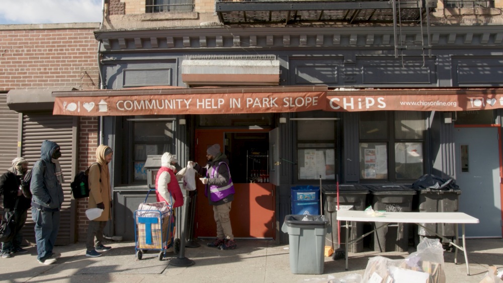 People standing outside a community help center, CHIP in Park Slope, with a sign above. One person wears a purple jacket giving a bag to another with carts nearby.