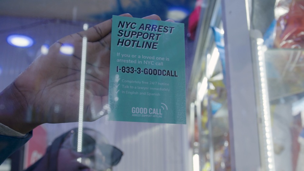 A person holds a green flyer against a window. The flyer advertises the NYC Arrest Support Hotline with a phone number and mentions 24/7 free support in English and Spanish.