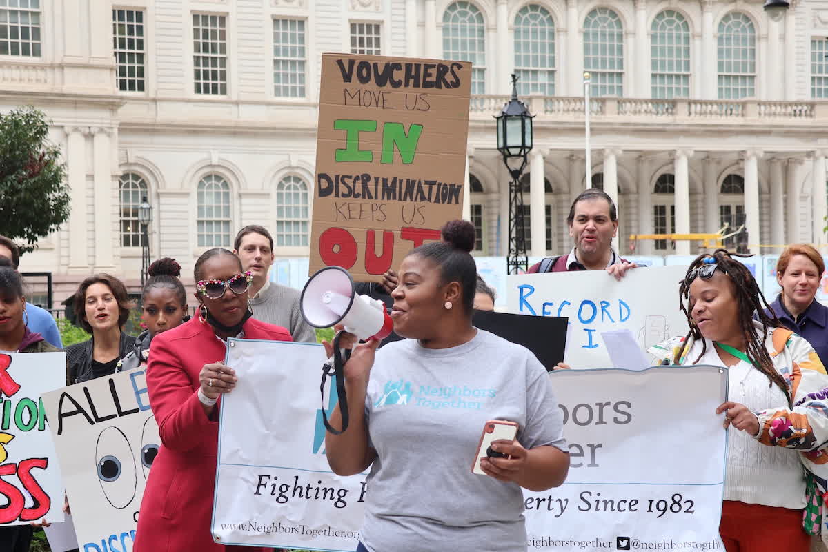 A group of people protest outside a building. One person holds a megaphone, while another holds a sign that reads, "Vouchers Move Us In, Discrimination Keeps Us Out.