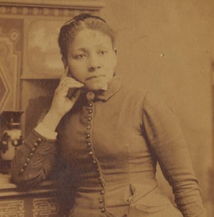 A woman in 19th-century attire poses with her hand on her chin, standing next to an ornately decorated piece of furniture.