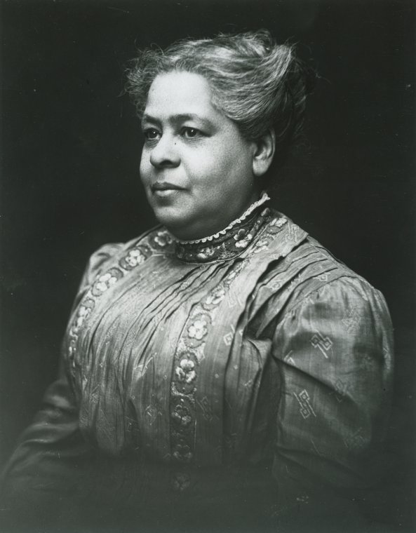 A woman with an elaborate updo hairstyle, wearing a detailed, high-collared dress with embroidered decorative patterns, poses in a seated position against a dark background.