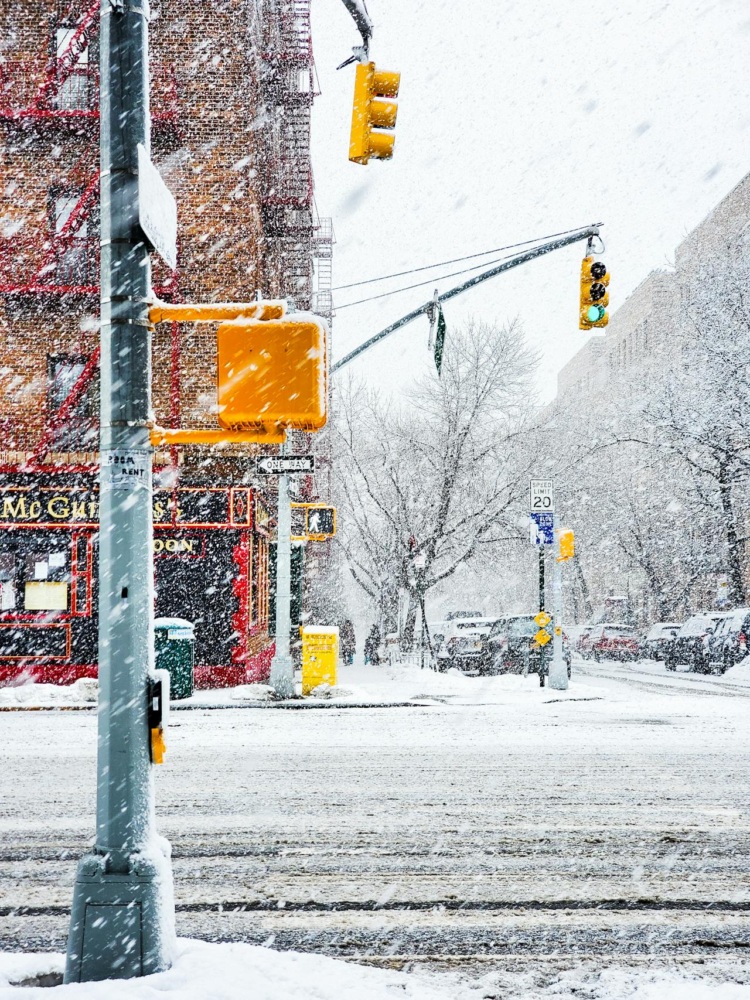 A snowy urban street intersection with traffic lights and cars. Snow covers the road and nearby buildings, creating a wintry scene. Red neon lights are visible on a building.