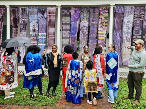 A group of people stand outdoors in front of a wall with colorful textiles. Some wear bright, patterned coats. One person holds an umbrella, and another gestures with a fist.