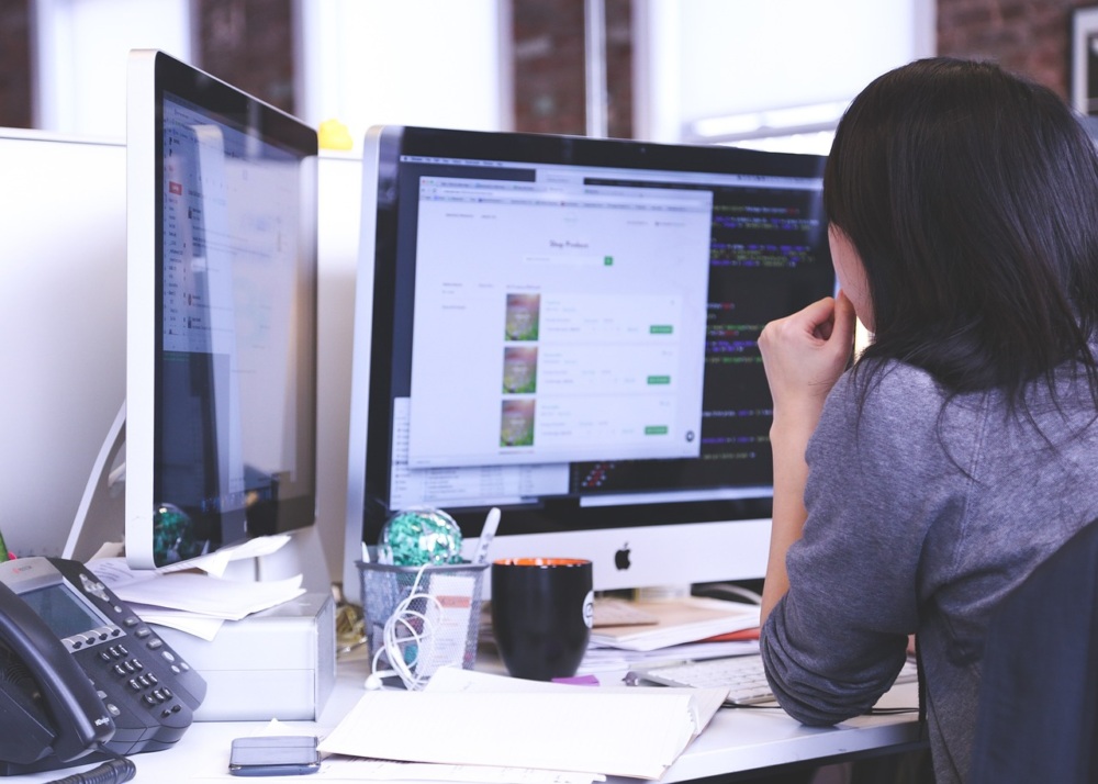 Person working at a desk with dual monitors, a notebook, phone, and a coffee mug, viewing web pages and code.