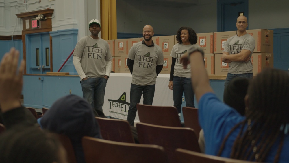 Group of four people wearing matching "Tech F.L.N." shirts stand on a stage in front of an audience, with boxes stacked behind them.