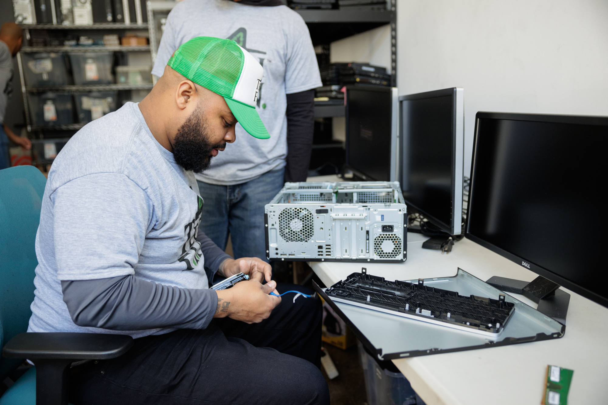 A person wearing a green cap and grey T-shirt works on assembling a computer at a desk, with open computer cases and monitors nearby.