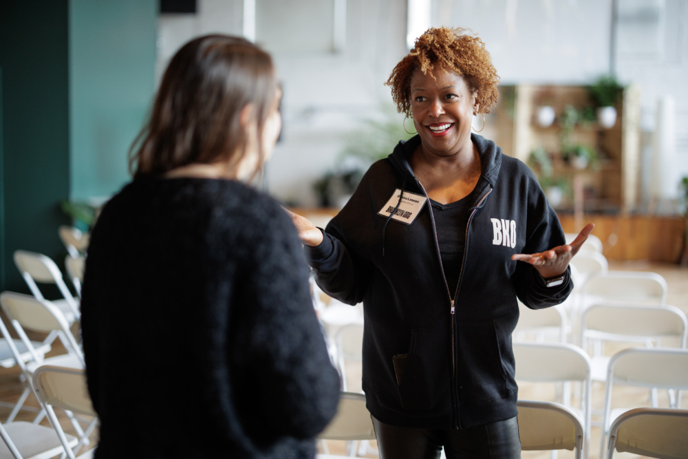 Two people in conversation in a room with rows of empty chairs. One, facing forward, gestures with both hands and wears a black hoodie with "BKN" printed on it.