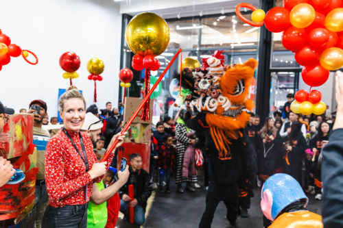 A woman dressed in red holds a stick with a red ribbon, standing near performers in a lion costume during a festive indoor celebration. Red and gold decorations are displayed overhead.