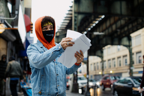 Person in orange hoodie and mask holds and points at papers on a city street under an elevated train track.