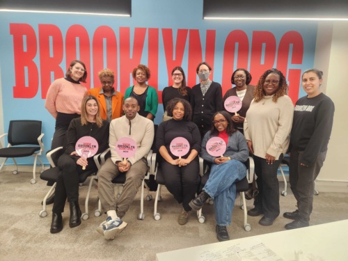 A group of people posing indoors, some seated and others standing, in front of a large "BROOKLYNORG" sign. Some hold fans with the same text.